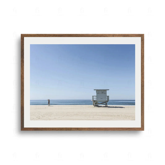 Plakat przedstawiający szeroką piaszczystą plażę, błękitny ocean, jasnoniebieską budkę ratownika z numerem 17 oraz sylwetkę osoby wpatrzonej w horyzont.